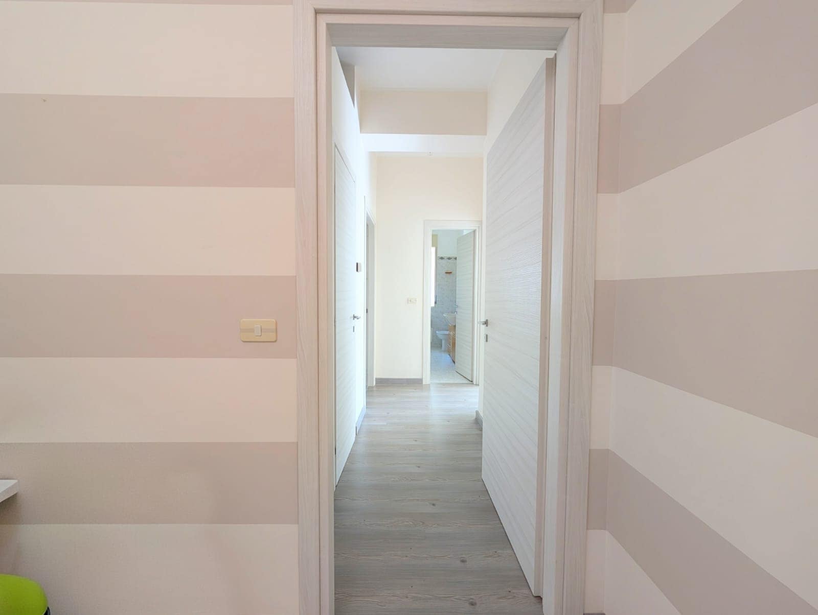 Ampio corridoio in un appartamento moderno con pareti a righe beige e bianche, pavimento in legno chiaro, porte bianche e illuminazione naturale, ideale per ambienti residenziali di alta qualità.