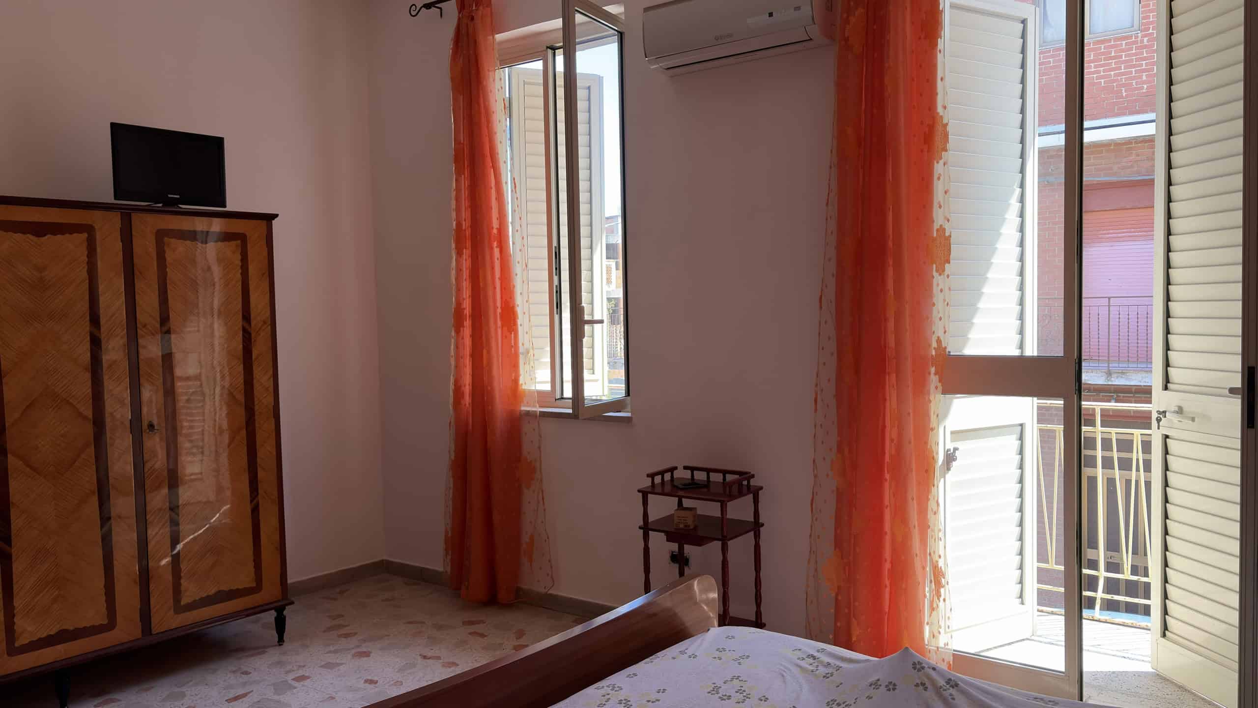 Ampia camera da letto con finestra e balcone, arredi eleganti e tende arancioni, ambiente luminoso e accogliente, ideale per soggiorno confortevole e relax.