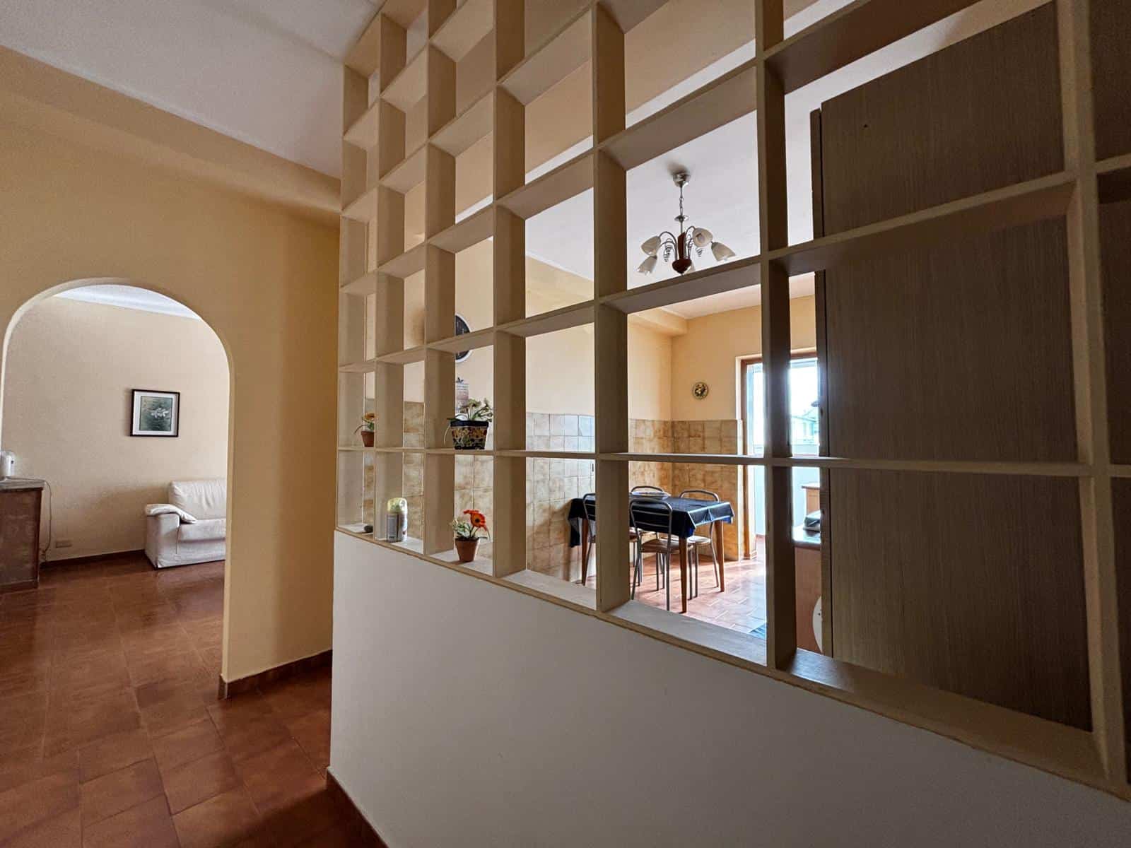 Interno di una casa con parete divisoria in legno a griglia, soggiorno luminoso con tavolo e sedie, pareti beige e pavimento in piastrelle marroni, stile classico e accogliente.