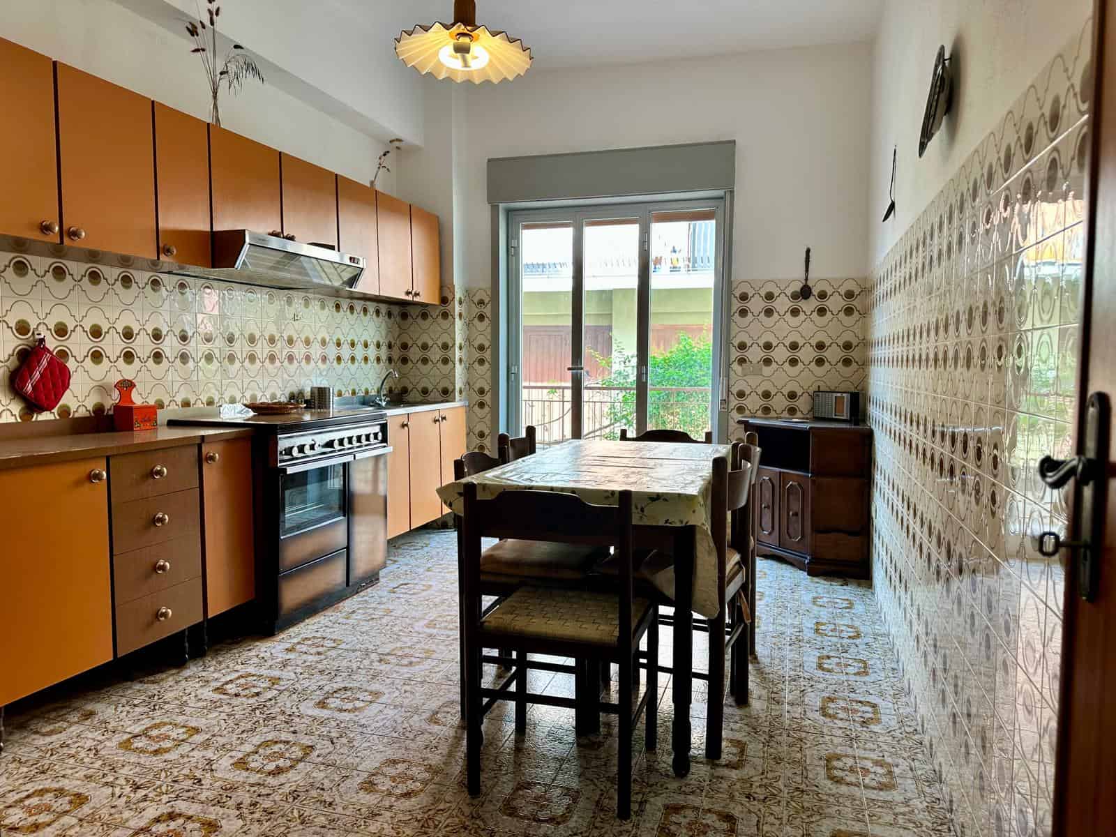 Cucina abitabile con mobili in legno, piano cottura, frigorifero e accessori, illuminata da luce naturale attraverso una porta finestra, con pareti rivestite in piastrelle decorative.
