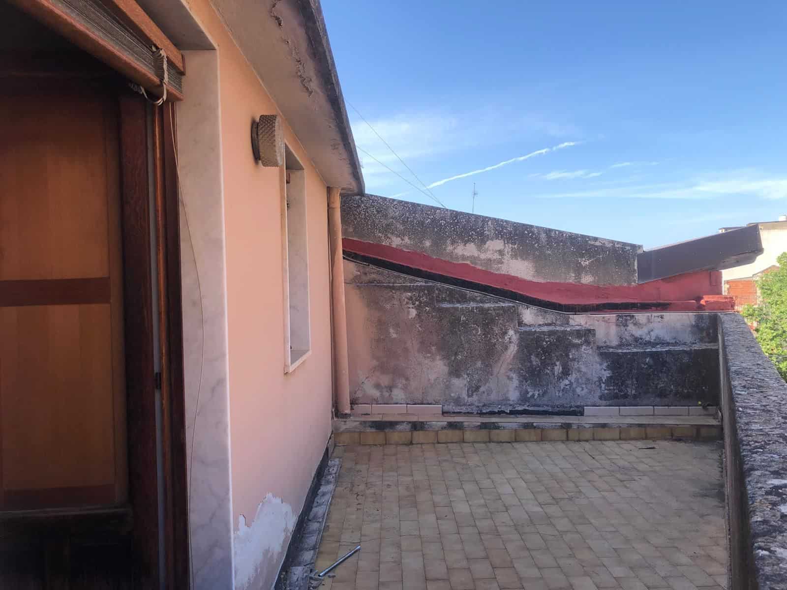 Balcone di un appartamento con pareti esterne in pietra e pavimento in piastrelle, vista sul cielo azzurro e su un edificio vicino, in un contesto urbano.