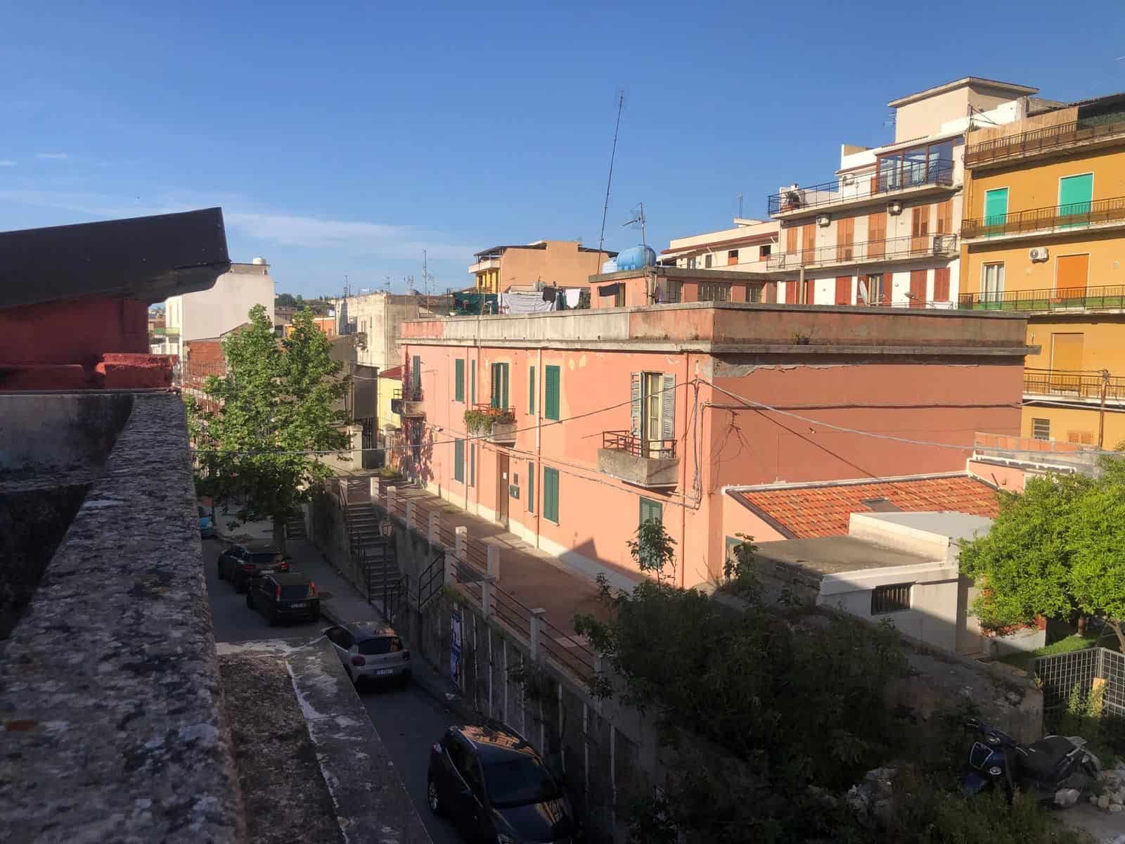 Balcone con vista su edifici residenziali colorati e strada tranquilla in un quartiere urbano di NextCasa, in Italia, con cielo sereno e vegetazione lussureggiante.