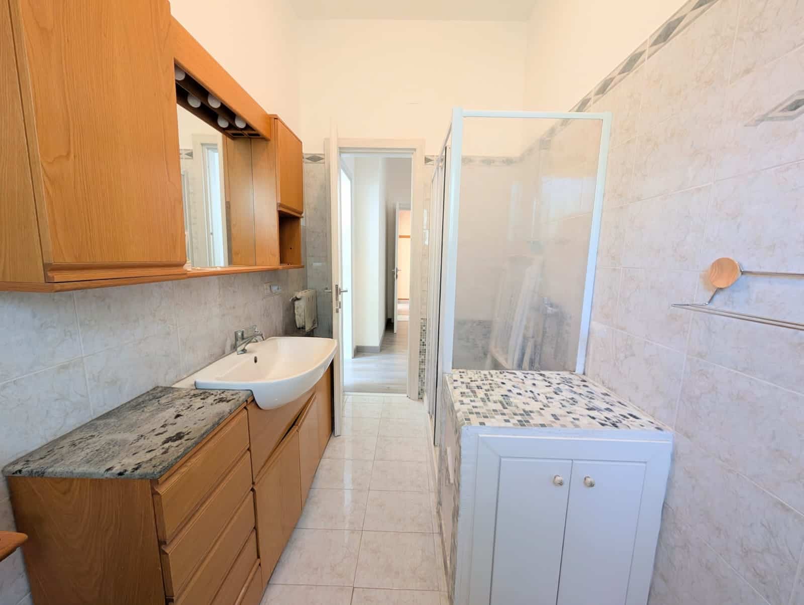 Lavabo e mobile bagno in legno con specchio e armadietto, bagno compatto e funzionale con pareti in piastrelle chiare e doccia separata, ideale per spazi piccoli e moderni.