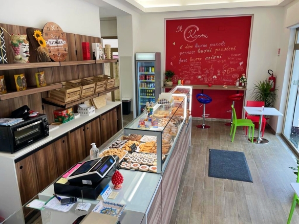 Panoramica di una caffetteria moderna con display di dolci e snack, pareti decorate con scritte motivazionali, area accogliente con sedie colorate e grande finestra che permette l'ingresso di luce nat.