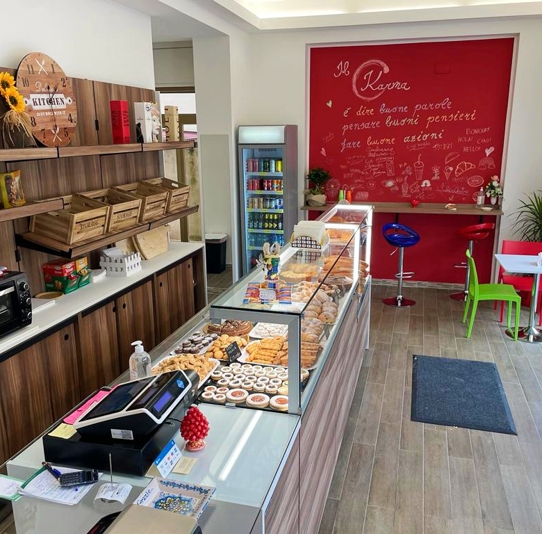 Panoramica di una caffetteria moderna con display di dolci e snack, pareti decorate con scritte motivazionali, area accogliente con sedie colorate e grande finestra che permette l'ingresso di luce nat.