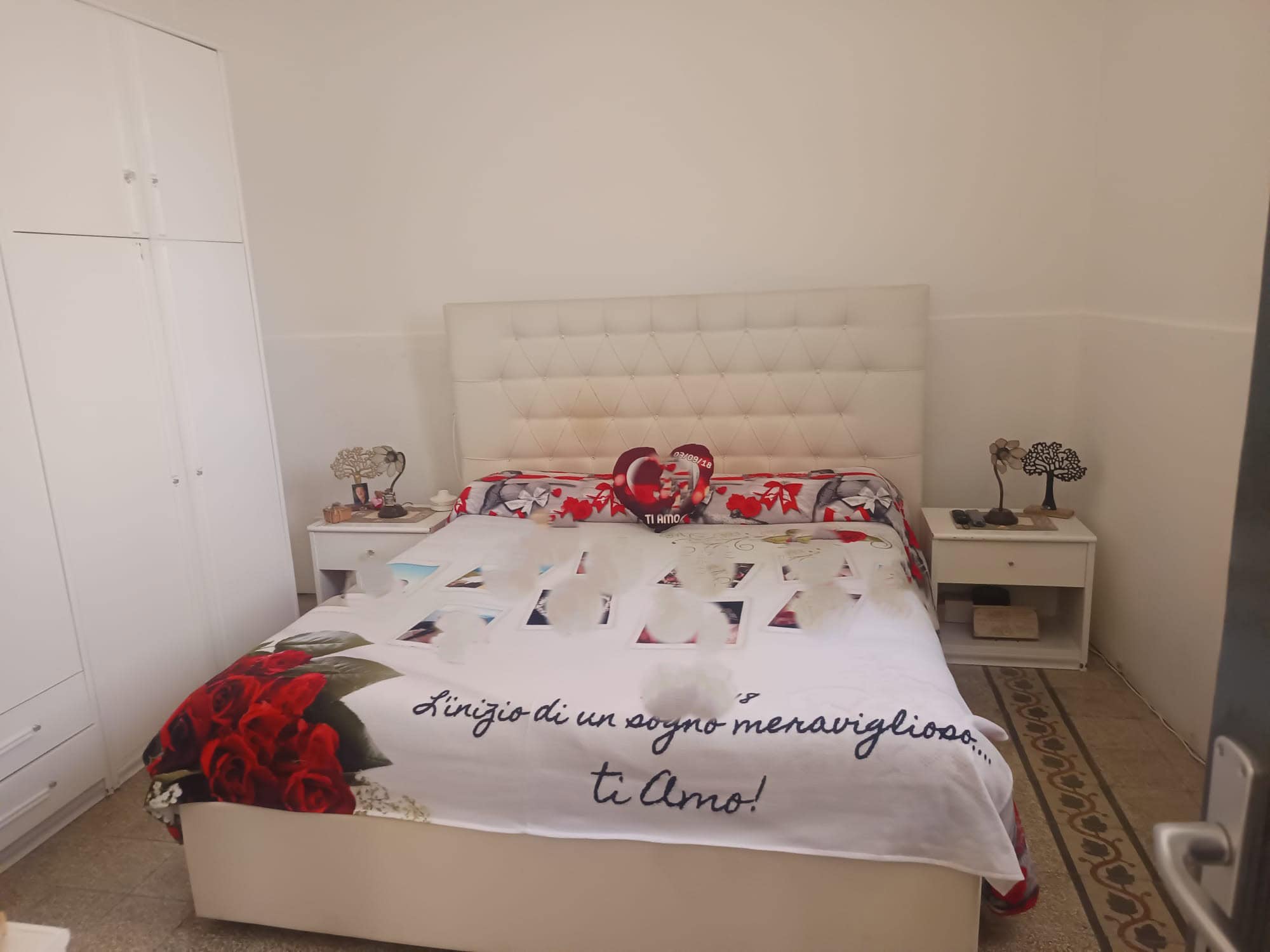 Un letto matrimoniale con testiera imbottita, coperta decorativa con scritta romantica e fiori rossi, in una camera da letto moderna e luminosa, con comodini e armadio bianchi.