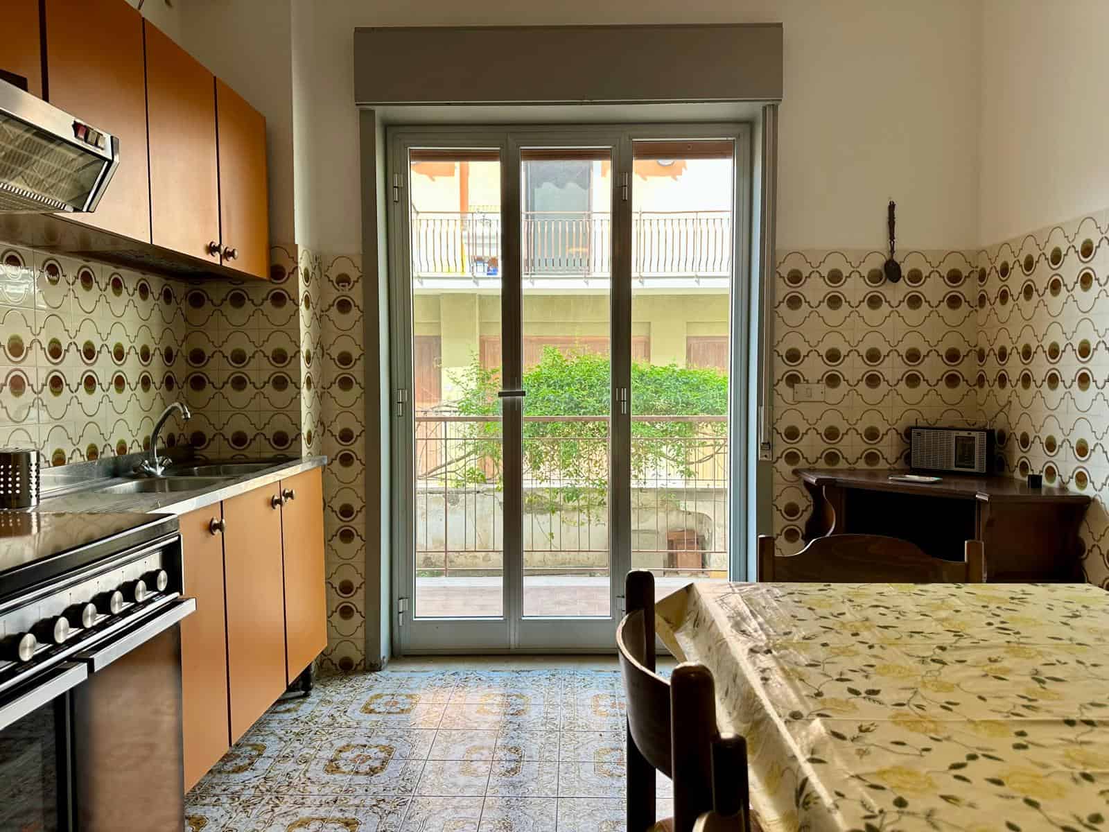 Porte scorrevoli in vetro che conducono a un balcone con vista su un giardino, in una cucina abitabile con mobili in legno, piastrelle decorative alle pareti e un tavolo con tovaglia floreale, ambient.