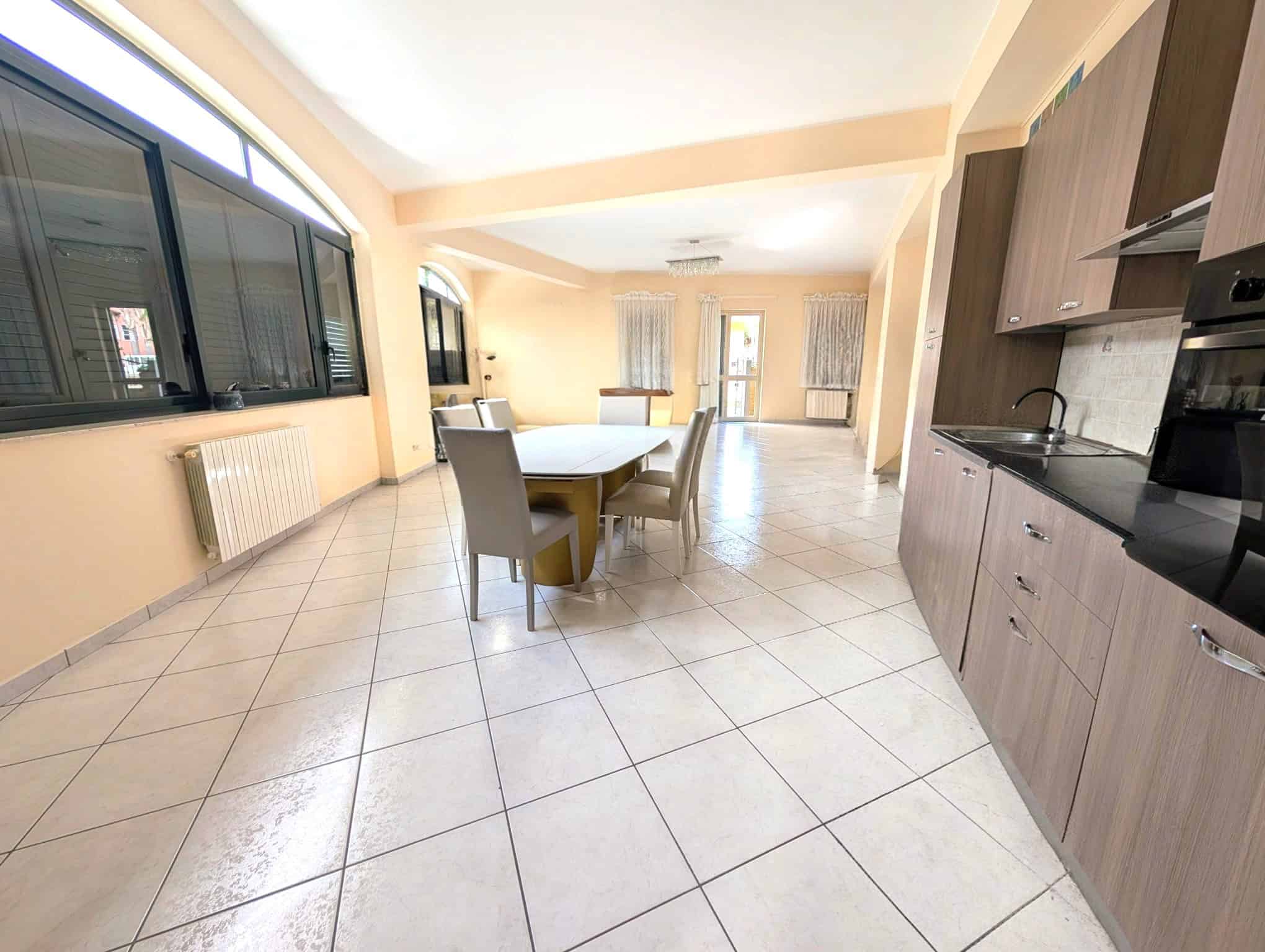 Ampia zona giorno open space con cucina moderna, tavolo da pranzo e grandi finestre che lasciano entrare molta luce naturale, ideale per vivere e ricevere ospiti in un ambiente confortevole e luminoso.