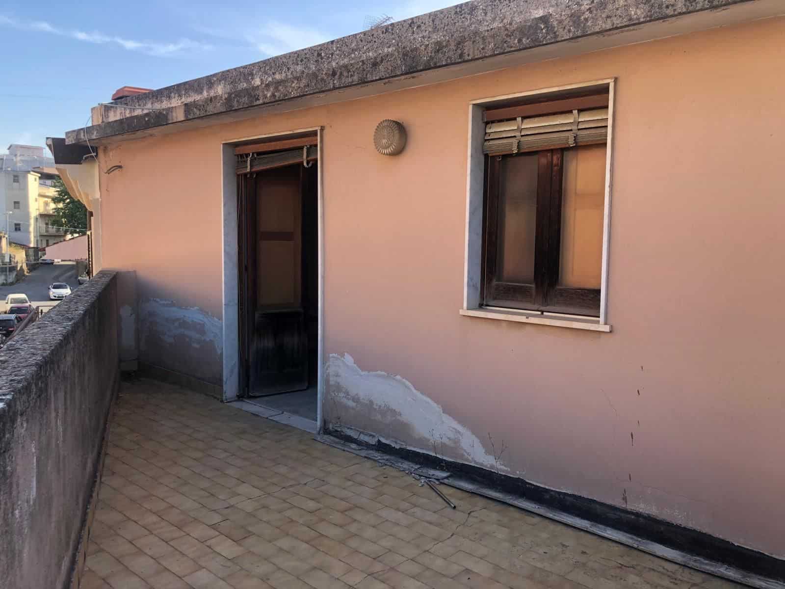 Balcone di un appartamento con pareti rosa, finestra e porta con tapparelle, pavimento in piastrelle beige, in un contesto urbano soleggiato.