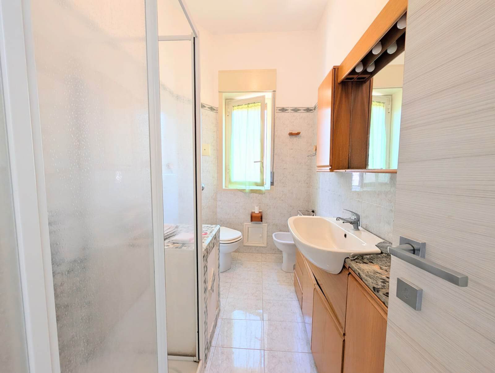 Lavabo e servizi igienici in un bagno luminoso e spazioso, con arredi in legno e pareti chiare, ideale per case vacanza o residenziali, con finestra che permette l'ingresso di luce naturale.