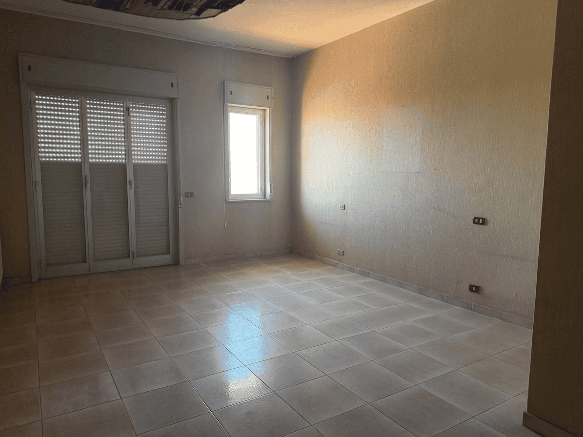 Stanza vuota con pareti beige, finestra e porta finestra con tapparelle, pavimento in piastrelle chiare, luce naturale, ideale per arredamento e ristrutturazione.