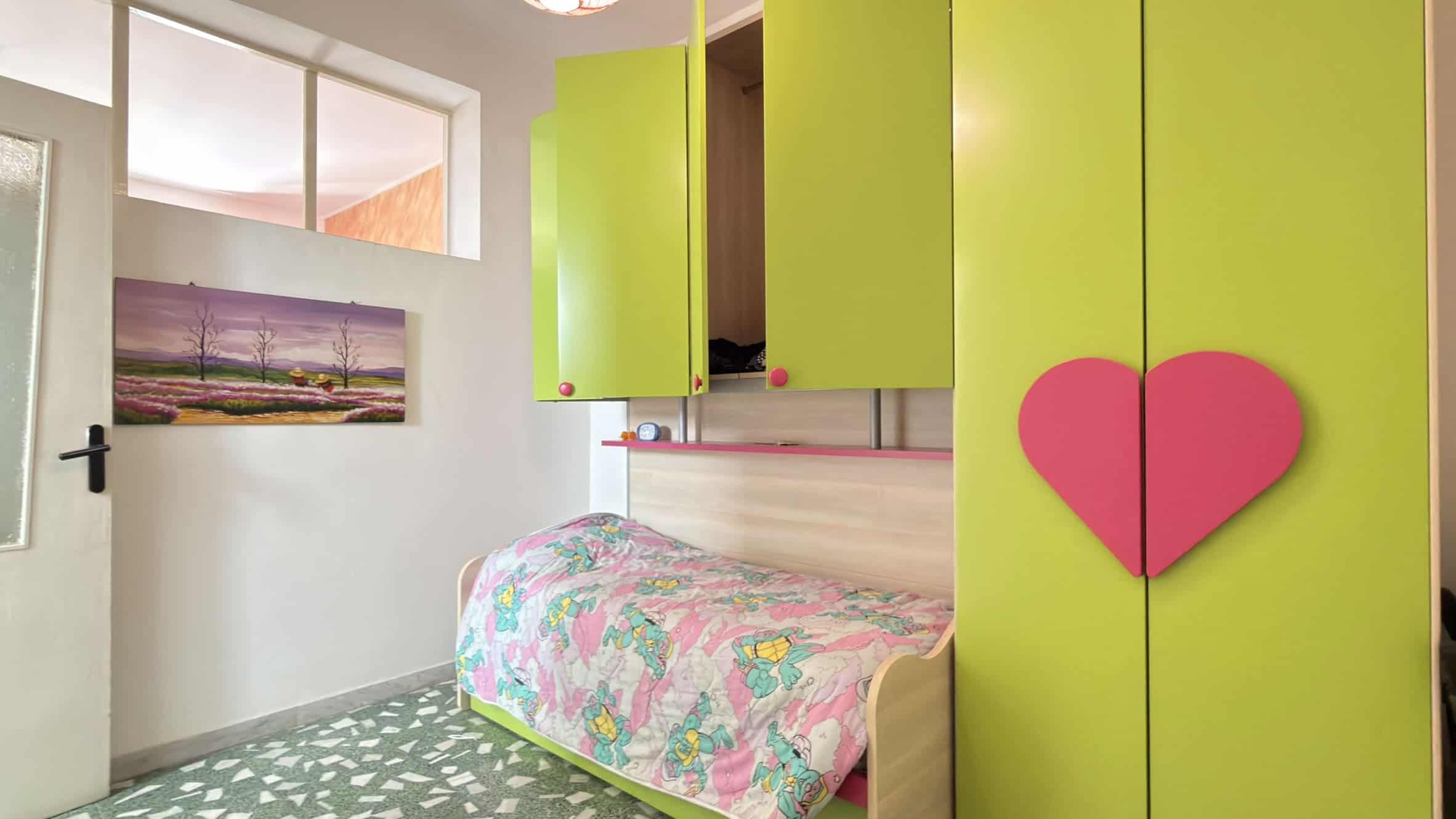 Letto singolo con coperta colorata in una stanza luminosa con pareti bianche, armadio verde brillante e decorazioni a forma di cuore rosa, ideale per ambienti giovani e vivaci.