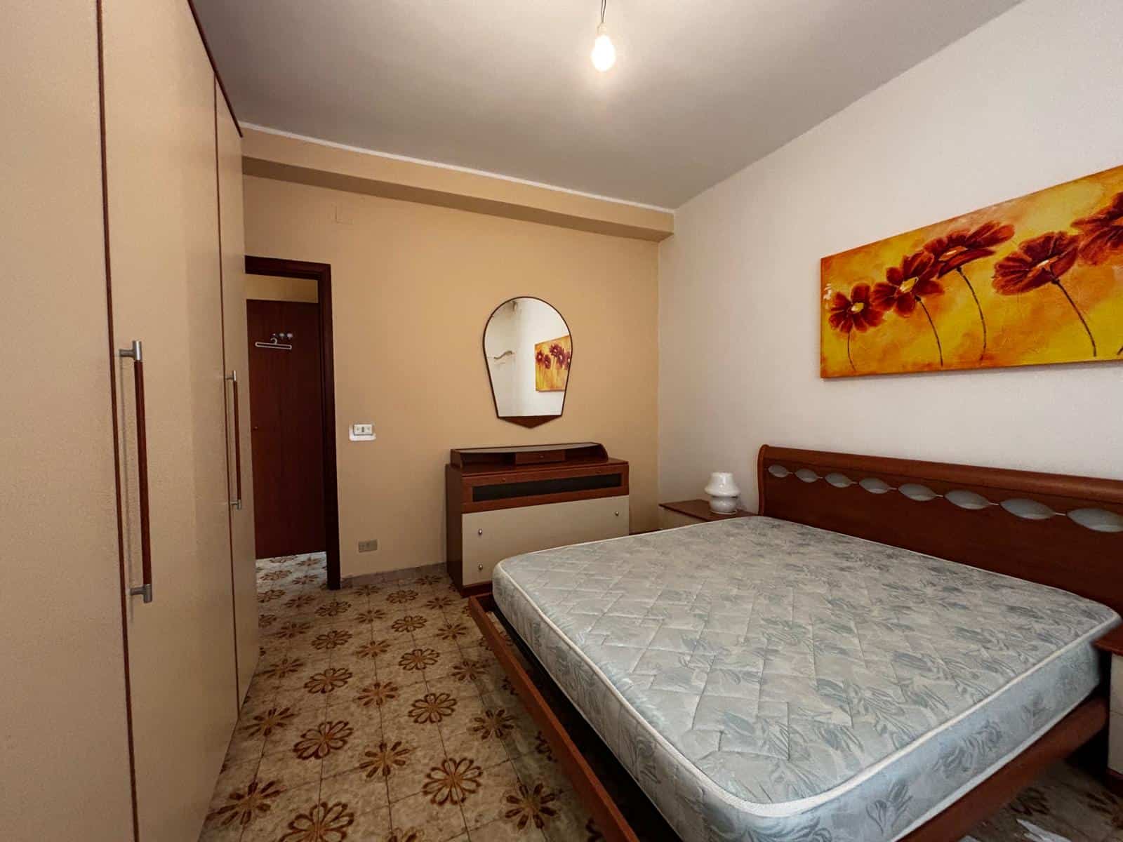 Letto matrimoniale con testiera in legno, armadio a muro e mobiletto con specchio in una camera da letto luminosa e accogliente. Ideale per appartamenti moderni e confortevoli.