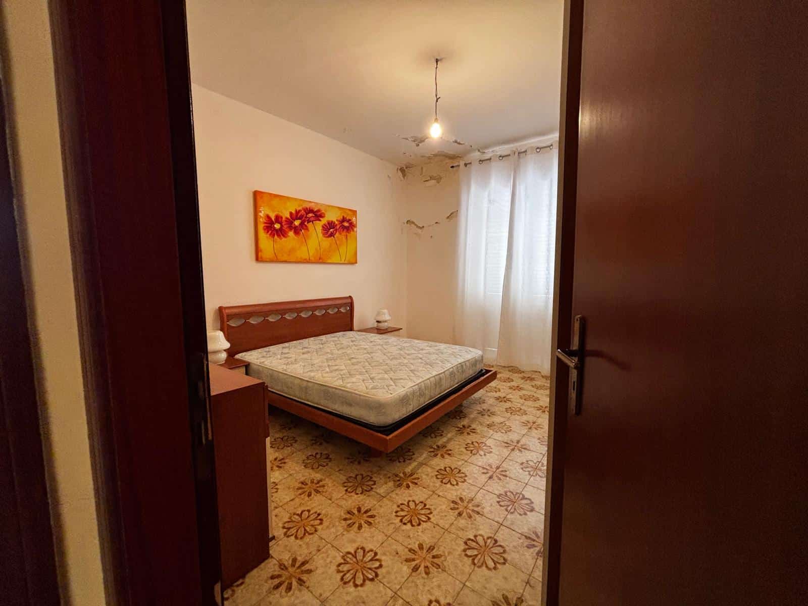 Il letto matrimoniale in una camera da letto semplice con pareti bianche, un quadro floreale sopra la testata e una finestra con tende leggere, in un ambiente accogliente e luminoso.