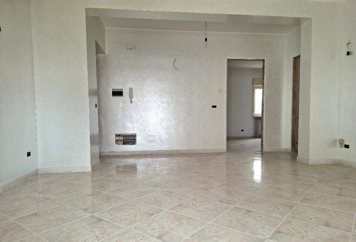 Appartamento in fase di ristrutturazione con pareti bianche e pavimento in gres chiaro, ampio spazio living con ingresso aperto e luminoso, pronto per personalizzazione e arredo.