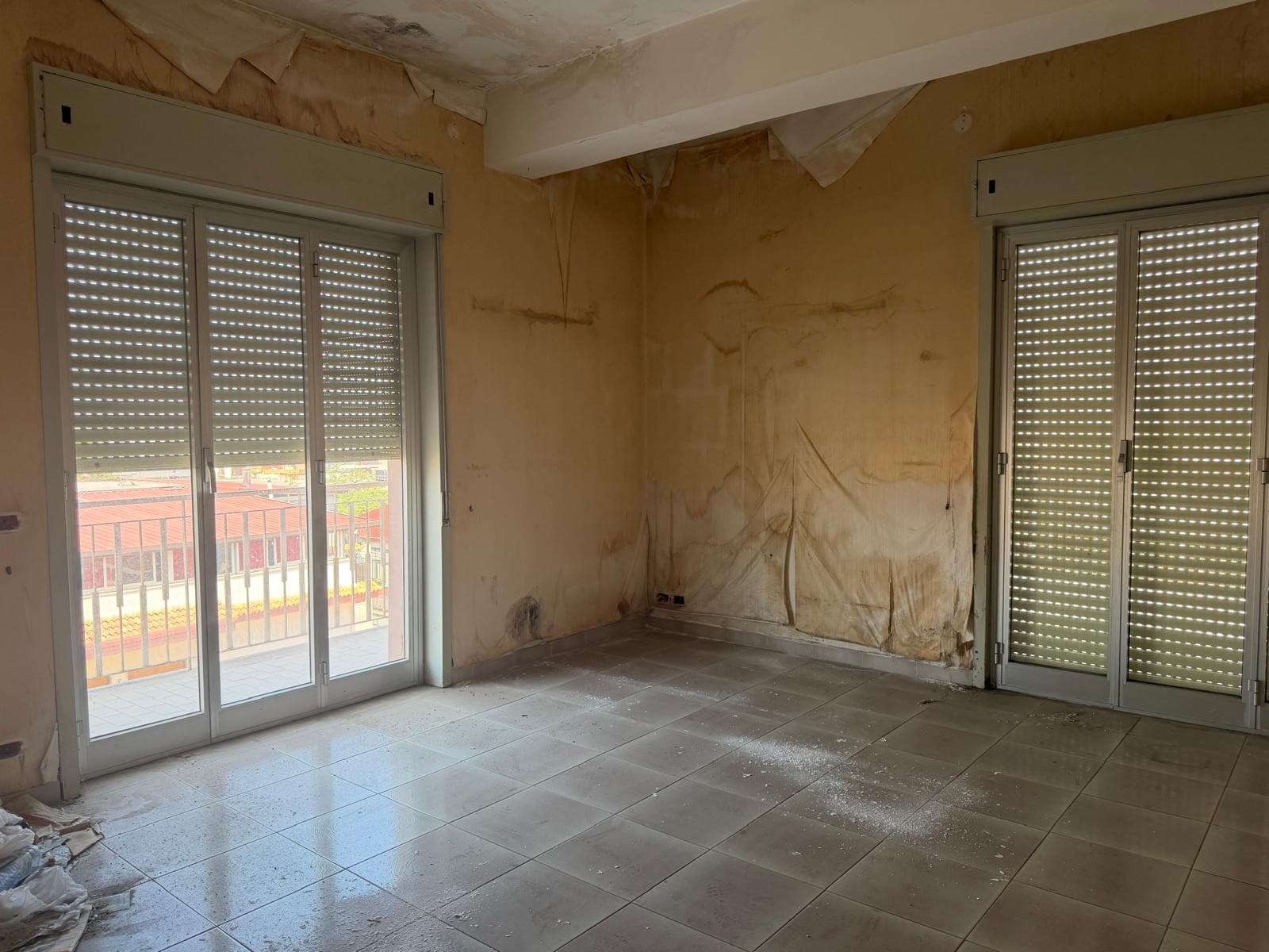 Serrande avvolgibili in alluminio su grandi finestre in un appartamento in fase di ristrutturazione, con pareti da rifinire e pavimento in piastrelle. Ideale per sicurezza e isolamento termico.