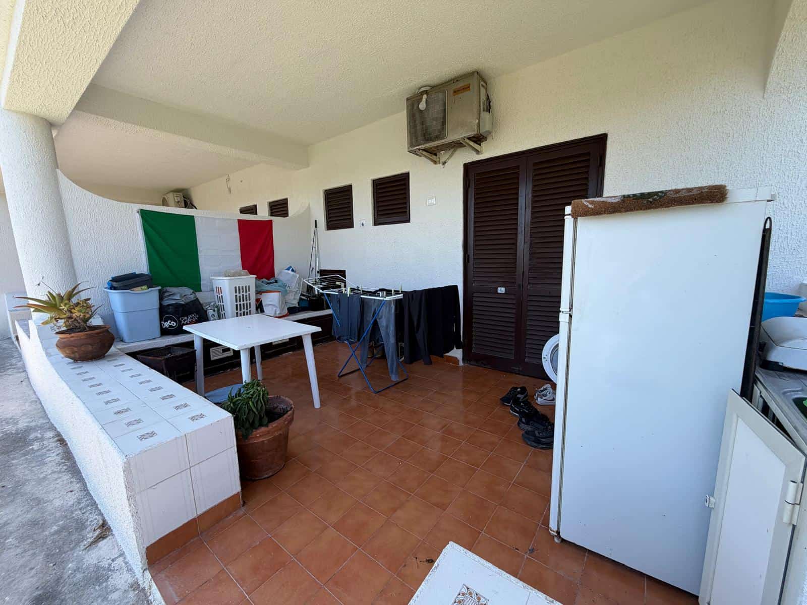 Balcone di un appartamento con piante, tavolo, stendibiancheria e bandiera italiana, in un contesto residenziale.