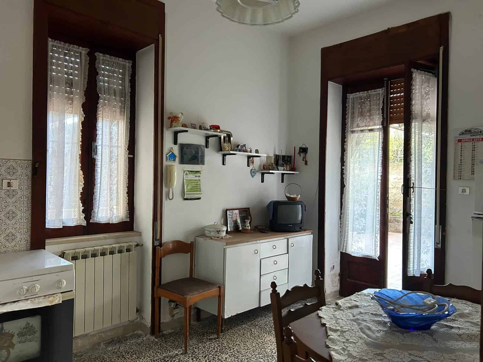 Interno di una casa con mobili vintage, finestra con tende di pizzo e porta finestra con persiane in legno, ambiente luminoso e accogliente.