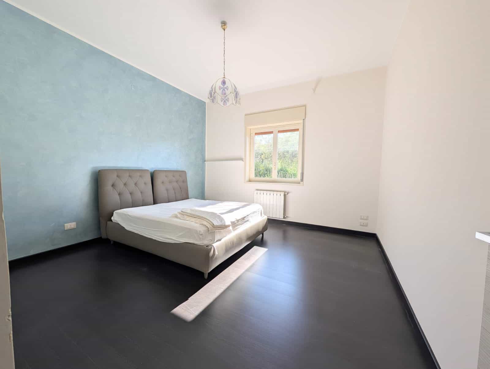 Letto matrimoniale elegante in camera da letto moderna con parete blu e pavimento scuro, luce naturale e arredamento minimalista.