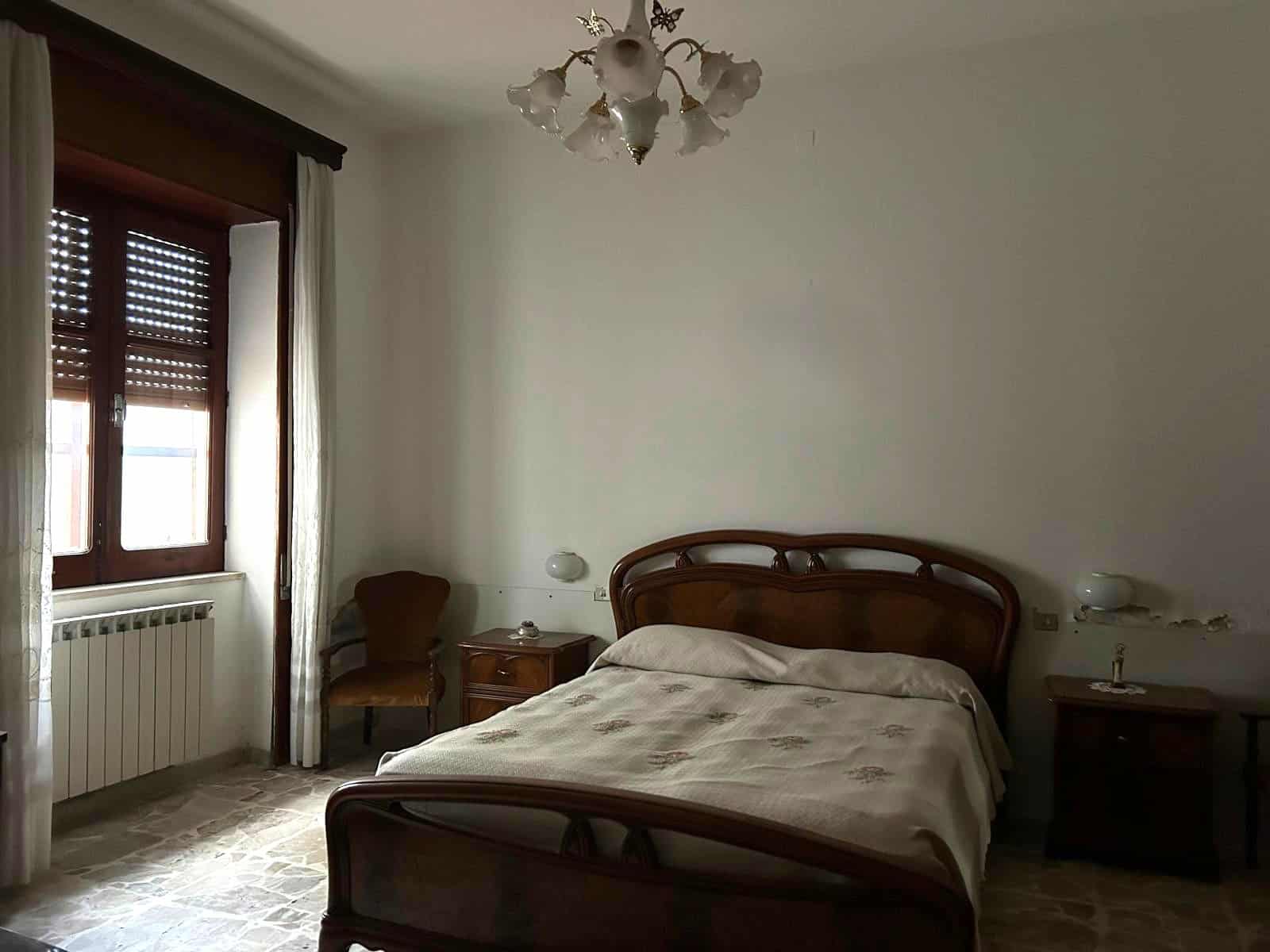 Letto matrimoniale in una stanza luminosa con pareti bianche, finestra con tende e arredi classici in legno, atmosfera tranquilla e accogliente, ideale per soggiorni rilassanti.