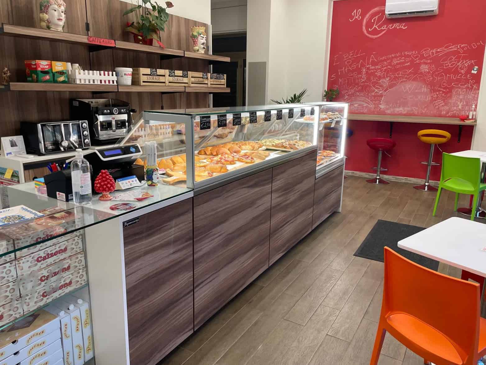 Pane di pizza e prodotti da forno all’interno di una vetrina refrigerata in una caffetteria o panetteria, con arredamento colorato e ambiente accogliente.