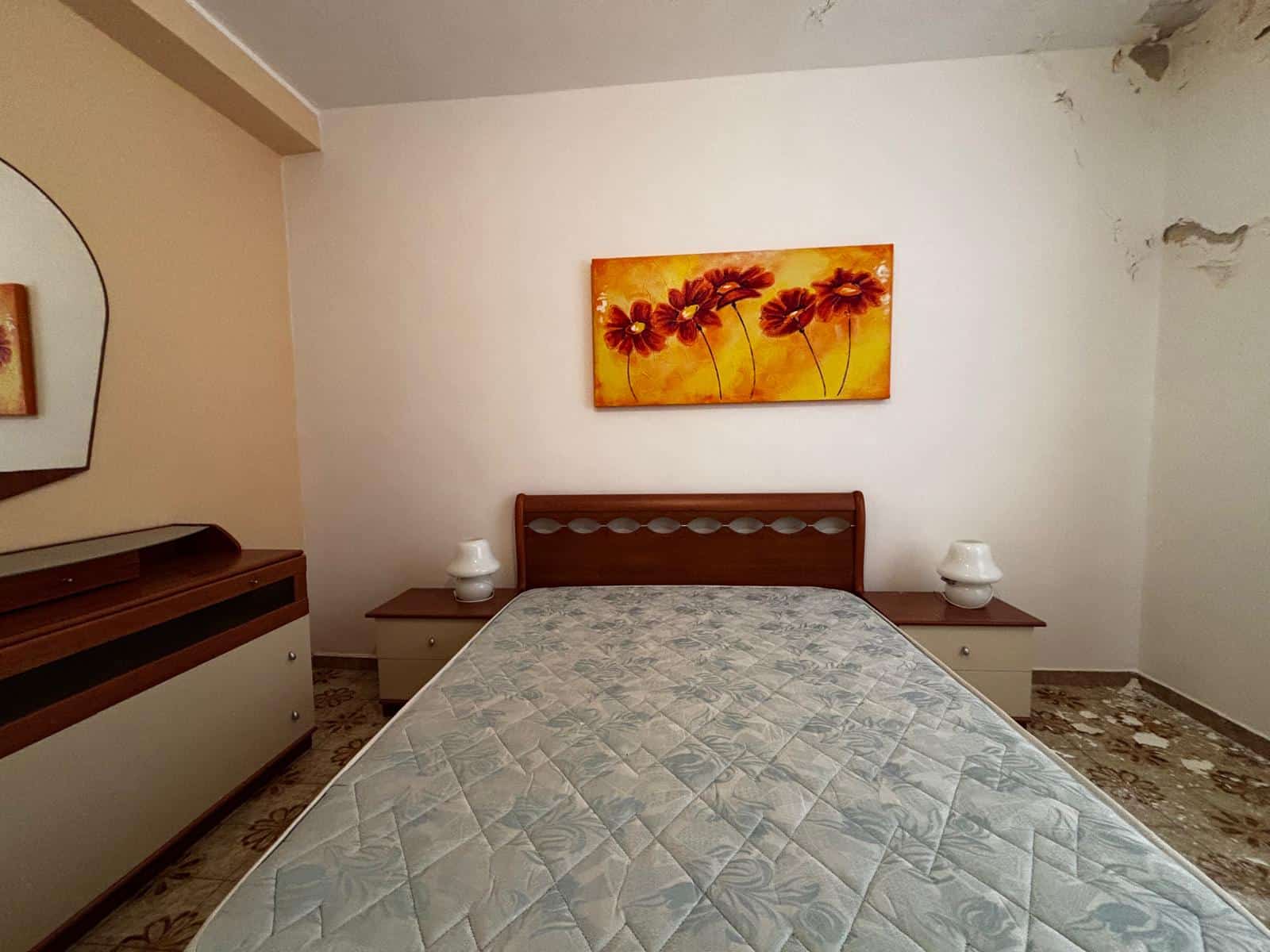 Il letto matrimoniale con testiera in legno, due comodini con lampade, e un quadro floreale arancione sopra il letto in una stanza semplice e luminosa.