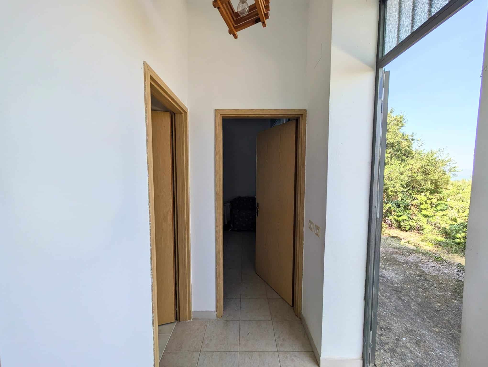 Ampia vista di un ingresso interno di una casa con pareti bianche e porte in legno, illuminata dalla luce naturale proveniente da una grande finestra, con vista su un paesaggio verde e naturale.