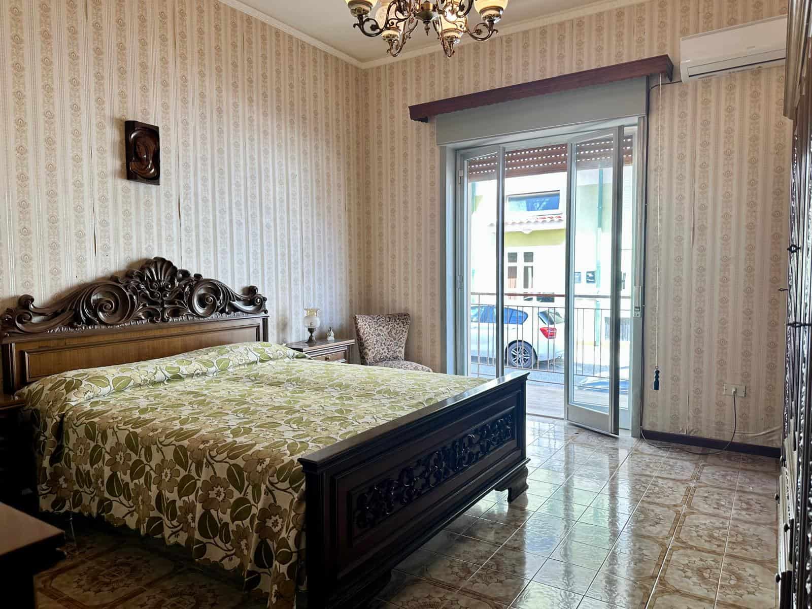 Elegante camera da letto con letto in legno intagliato, pareti rivestite in carta da parati beige, e accesso a un balcone con vista su strada e case circostanti, ideale per affitti turistici o residen.