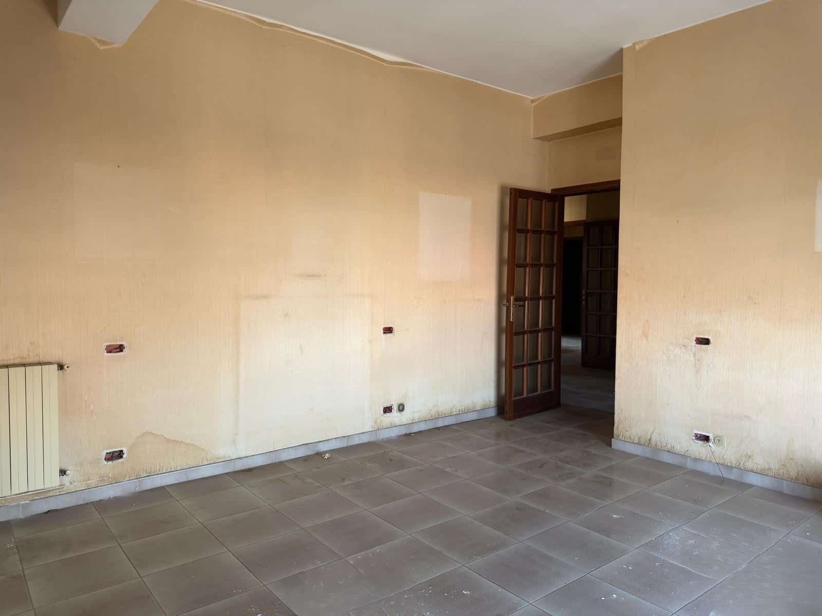 Interno di un appartamento vuoto con pareti beige, pavimento in piastrelle grigie e porta in legno, pronto per ristrutturazione o arredamento.