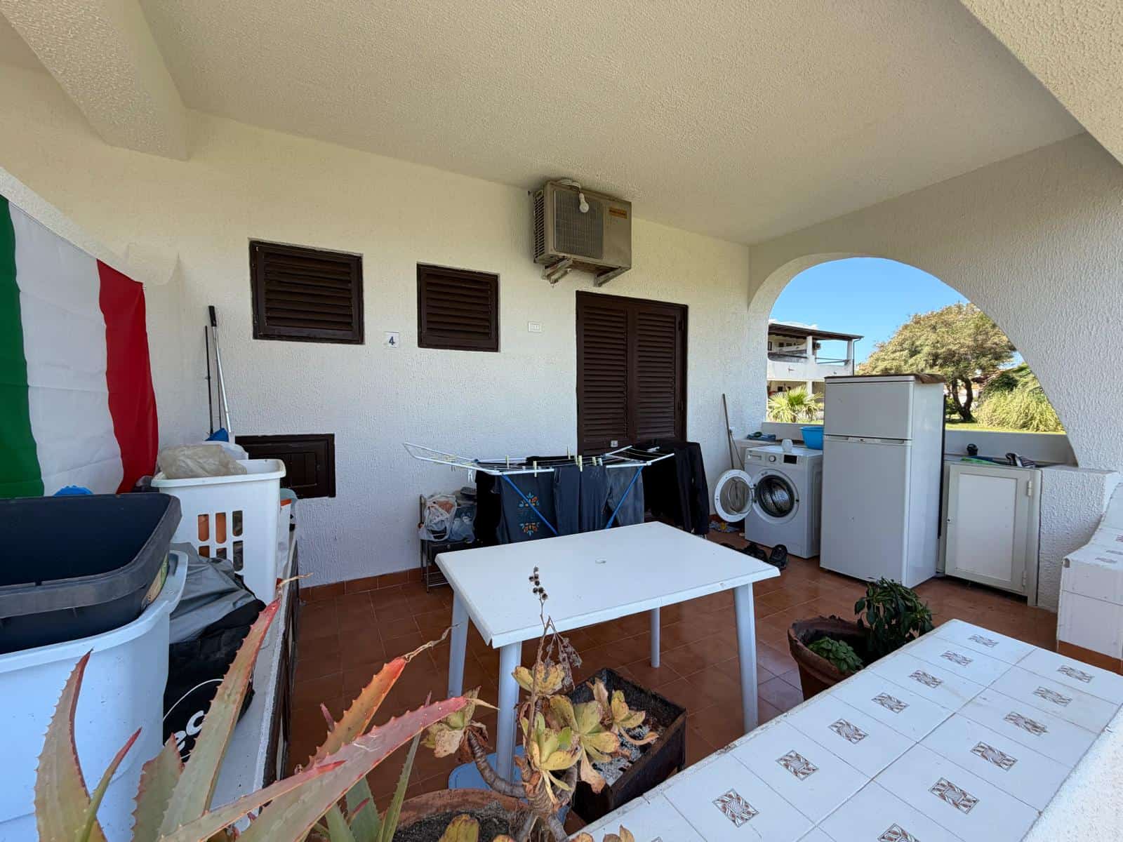 Lavanderia esterna con lavatrice, asciugatrice e spazio per il bucato, situata su un balcone coperto con vista sul giardino e sul cielo azzurro, ideale per il lavaggio e l'asciugatura all'aperto.