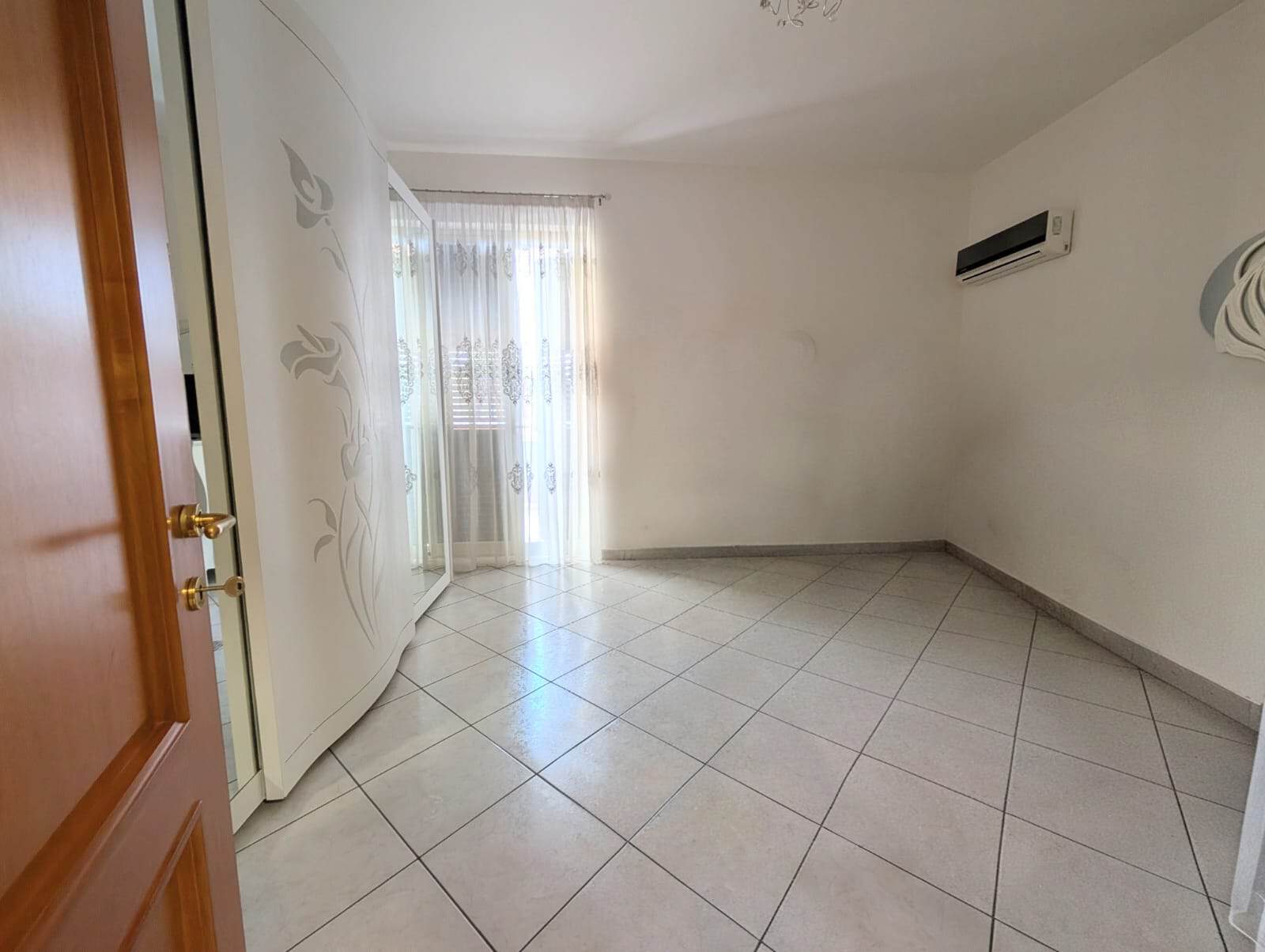 Stanza vuota con aria condizionata, pareti bianche e pavimento in piastrelle, luce naturale, ideale per affitto o vendita immobiliare.
