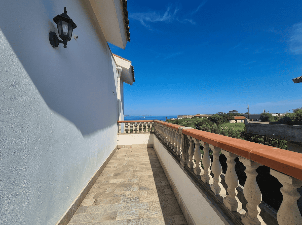 Ampia terrazza con vista panoramica sul mare, balaustra in stile classico, pavimentazione in piastrelle, parete bianca con lampada da esterno, cielo sereno e paesaggio verde in background.
