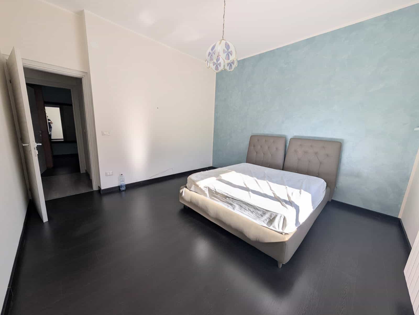 Letto matrimoniale in una camera moderna con parete blu e pavimento scuro, ambiente luminoso e minimalista, ideale per soggiorni confortevoli e rilassanti.