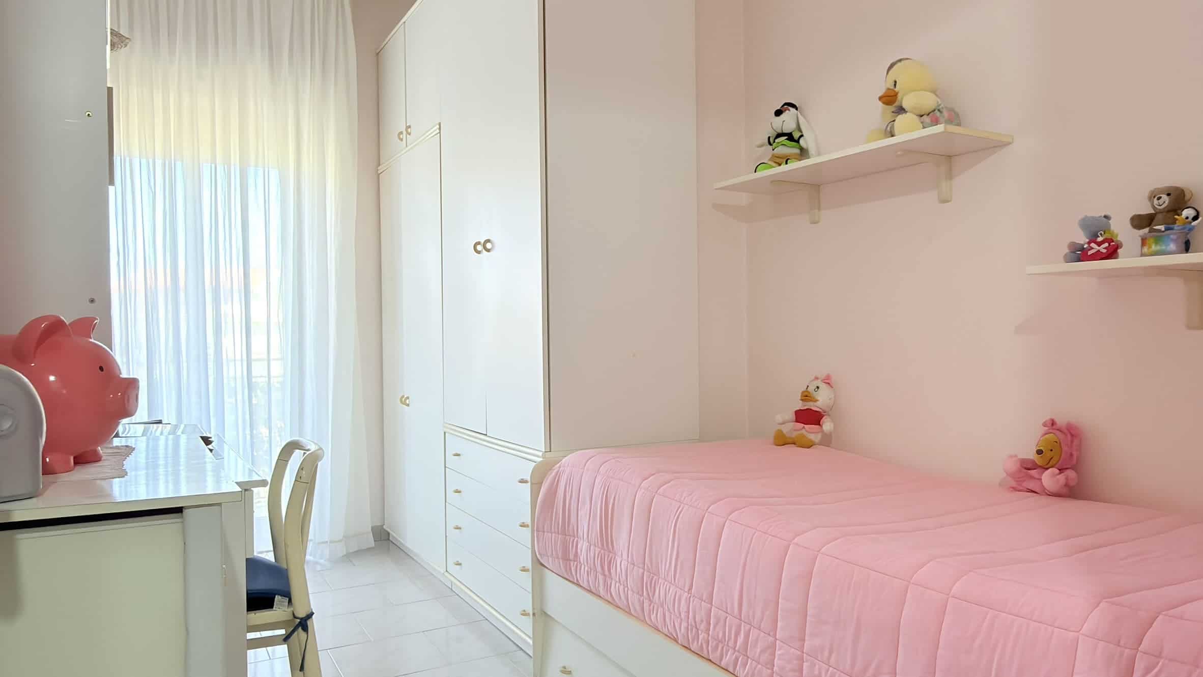 Ampio soggiorno con divano e zona pranzo, illuminato da grandi finestre con tende leggere, arredato con mobili bianchi e decorazioni colorate, ideale per vivere momenti di relax e convivialità.