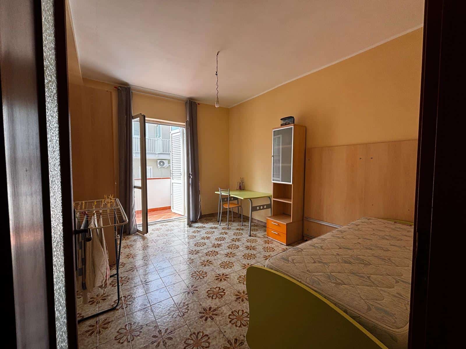 Letto matrimoniale in camera da letto con parete gialla, armadio e balcone con vista urbana, in un appartamento in affitto a NextCasa, Milano.