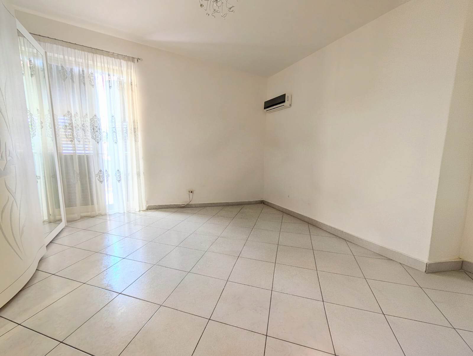 Ampia stanza luminosa con pareti bianche, pavimento in piastrelle e grande finestra con tende trasparenti, ideale per soggiorno o zona living.