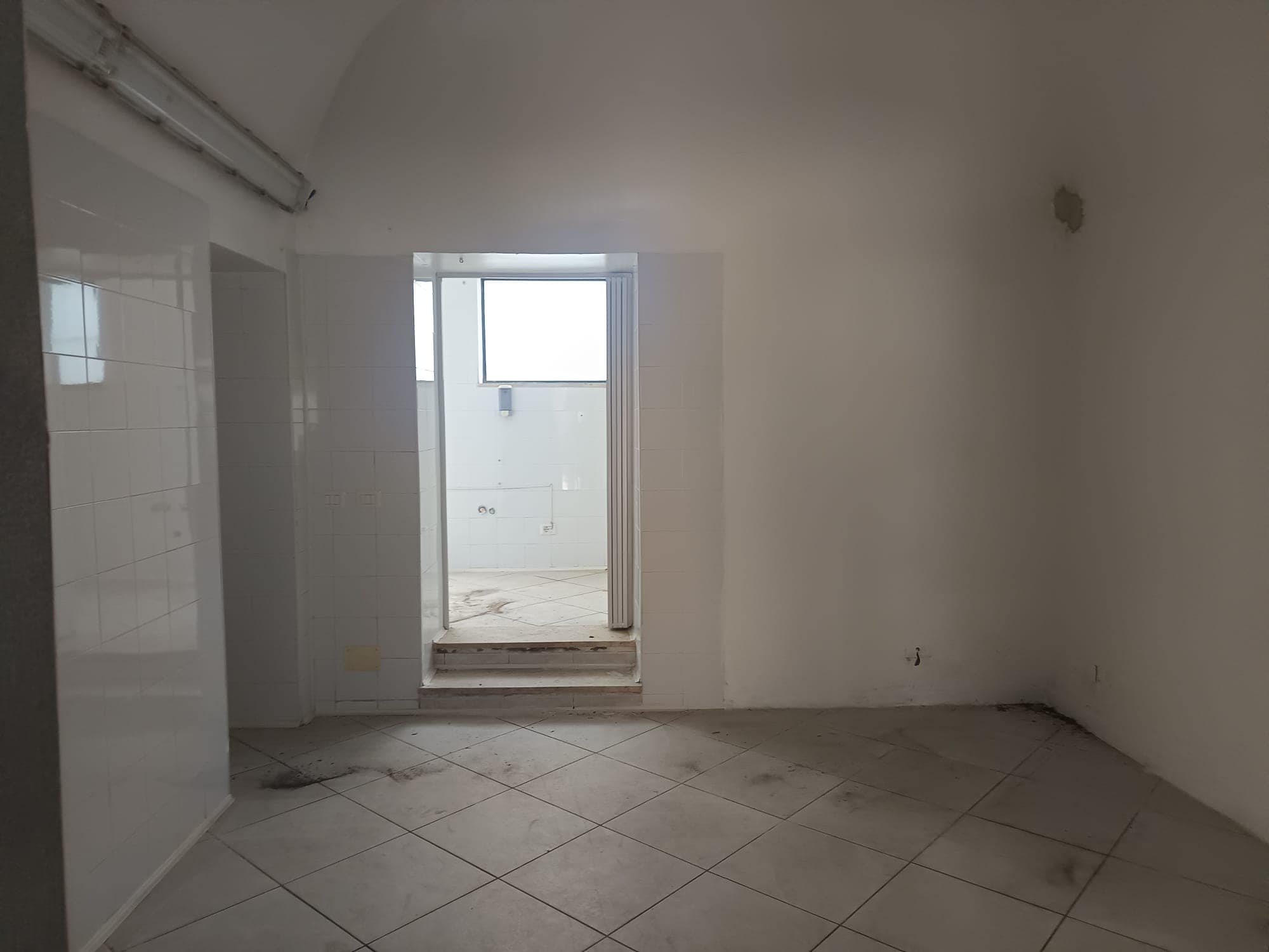 Ampio spazio vuoto in un appartamento in fase di ristrutturazione con pareti bianche e pavimento in piastrelle, luce naturale proveniente da una finestra e porta che conduce a un balcone o terrazza.