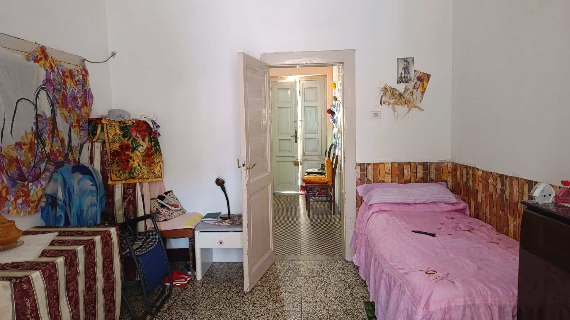 Una stanza da letto con pareti bianche, letto singolo con copriletto rosa, arredamento semplice e decorazioni colorate, in un ambiente domestico accogliente e luminoso.