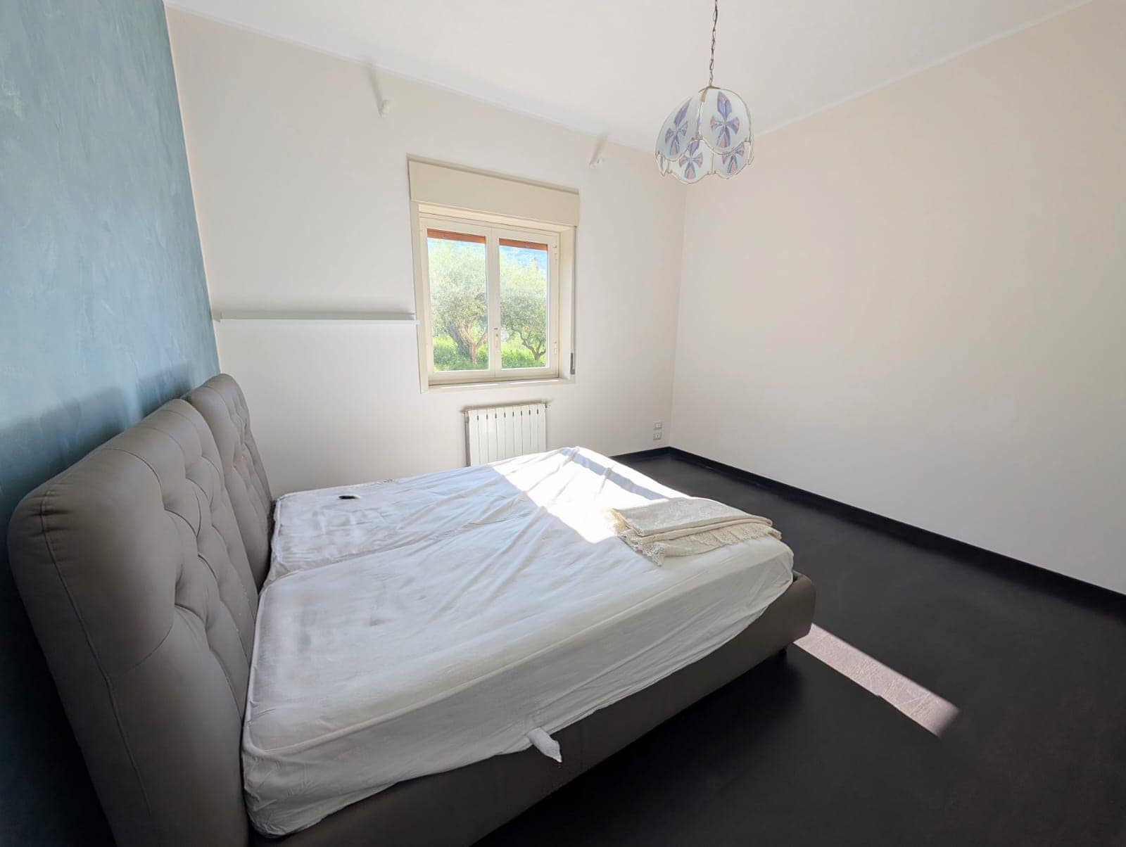 Aria aperta, camera da letto minimalista con letto matrimoniale, pareti bianche e finestra che lascia entrare luce naturale, pavimento scuro, decorazione semplice e atmosfera tranquilla.
