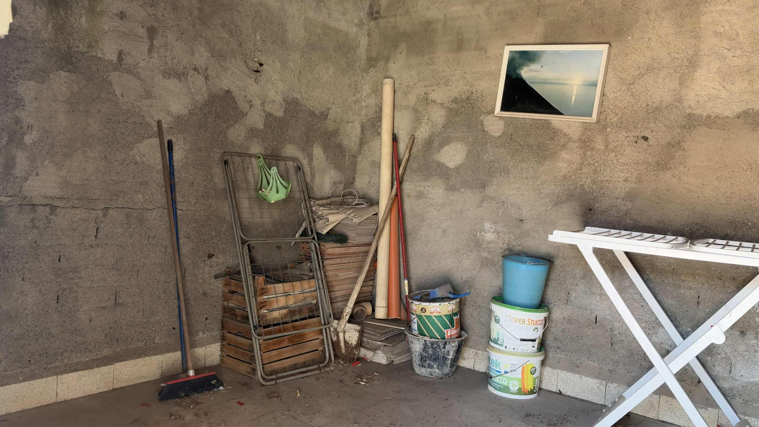 Spazzatura e attrezzi da giardinaggio in un angolo di un garage o cantina con pareti in cemento grezzo e pavimento in piastrelle.
