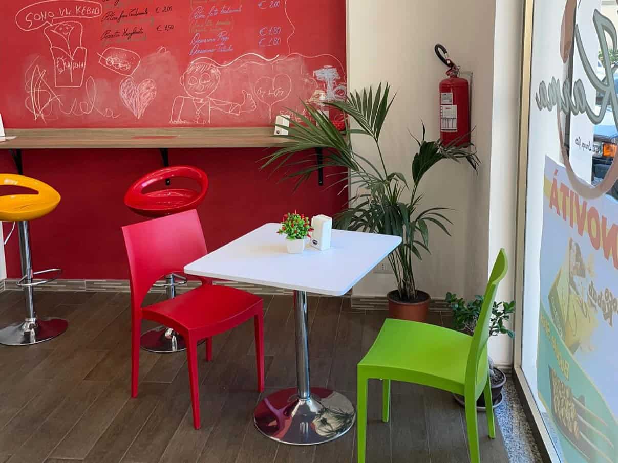 Un tavolo bianco con tre sedie colorate (rossa, verde e gialla) in un ambiente accogliente con pareti rosse e una lavagna con disegni e scritte.