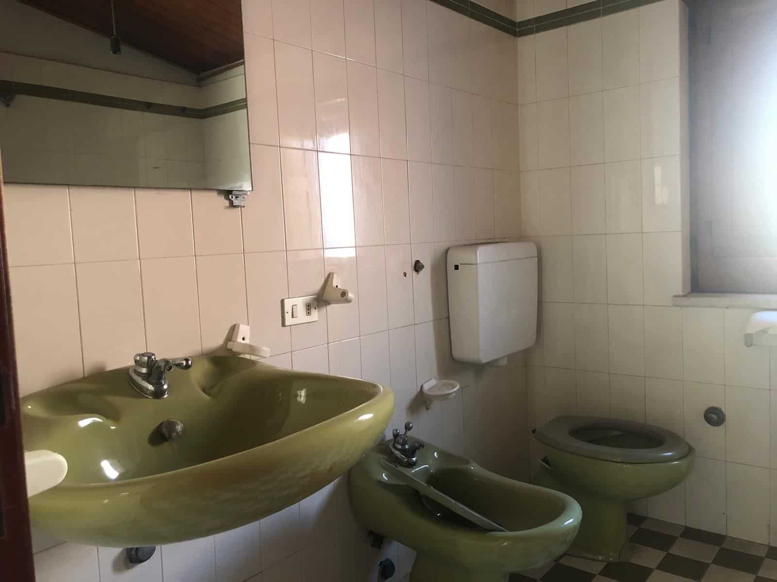 Lavabo e bidet verdi in un bagno con pareti rivestite di piastrelle beige e pavimento a scacchi. Ambiente semplice e funzionale, ideale per servizi igienici in abitazioni o strutture ricettive.