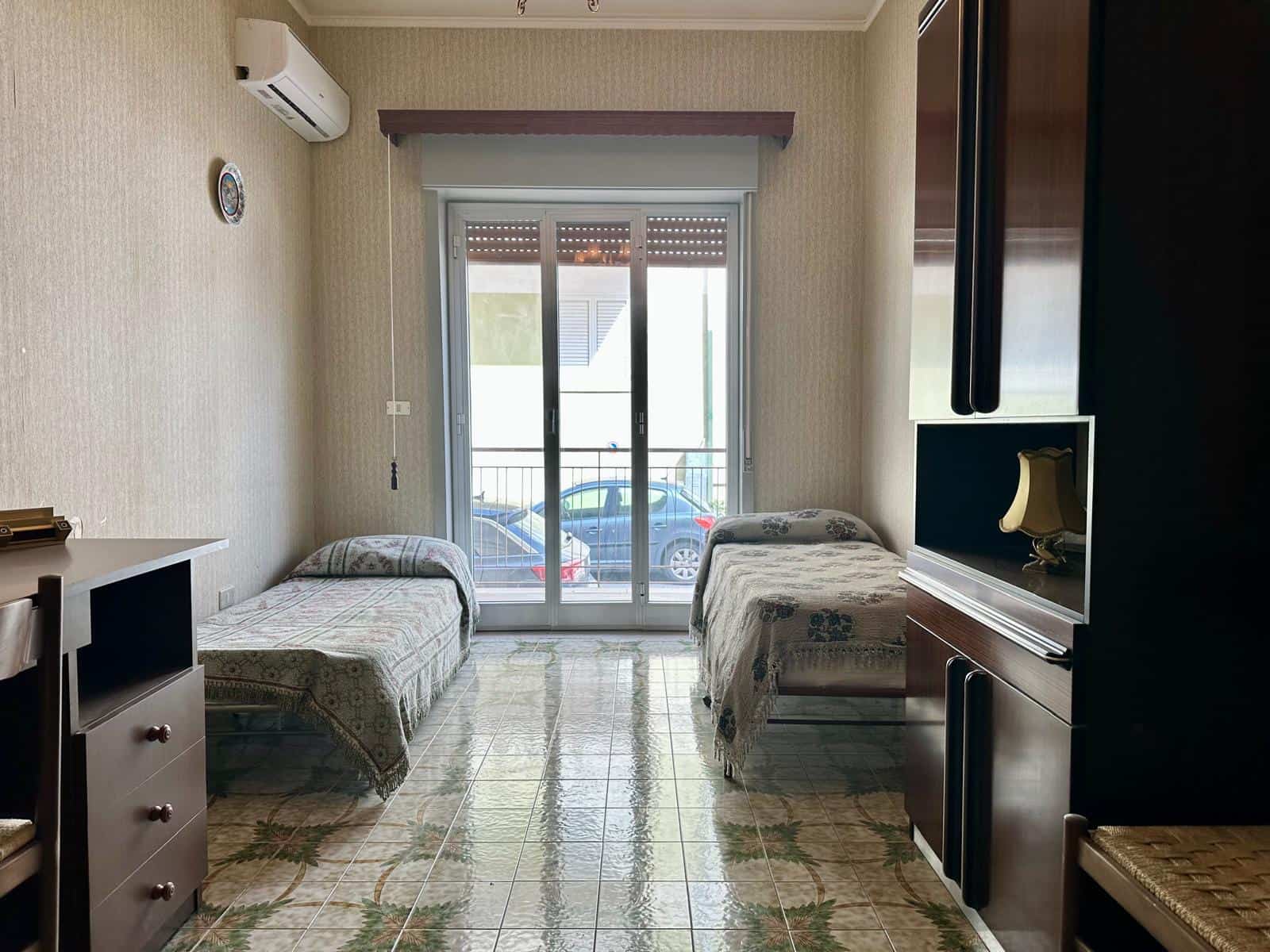 L'immagine mostra una camera da letto con due letti singoli, una grande finestra con porta scorrevole che dà su un balcone, e arredi moderni. Perfetta per affitti turistici o residenziali, con aria co.