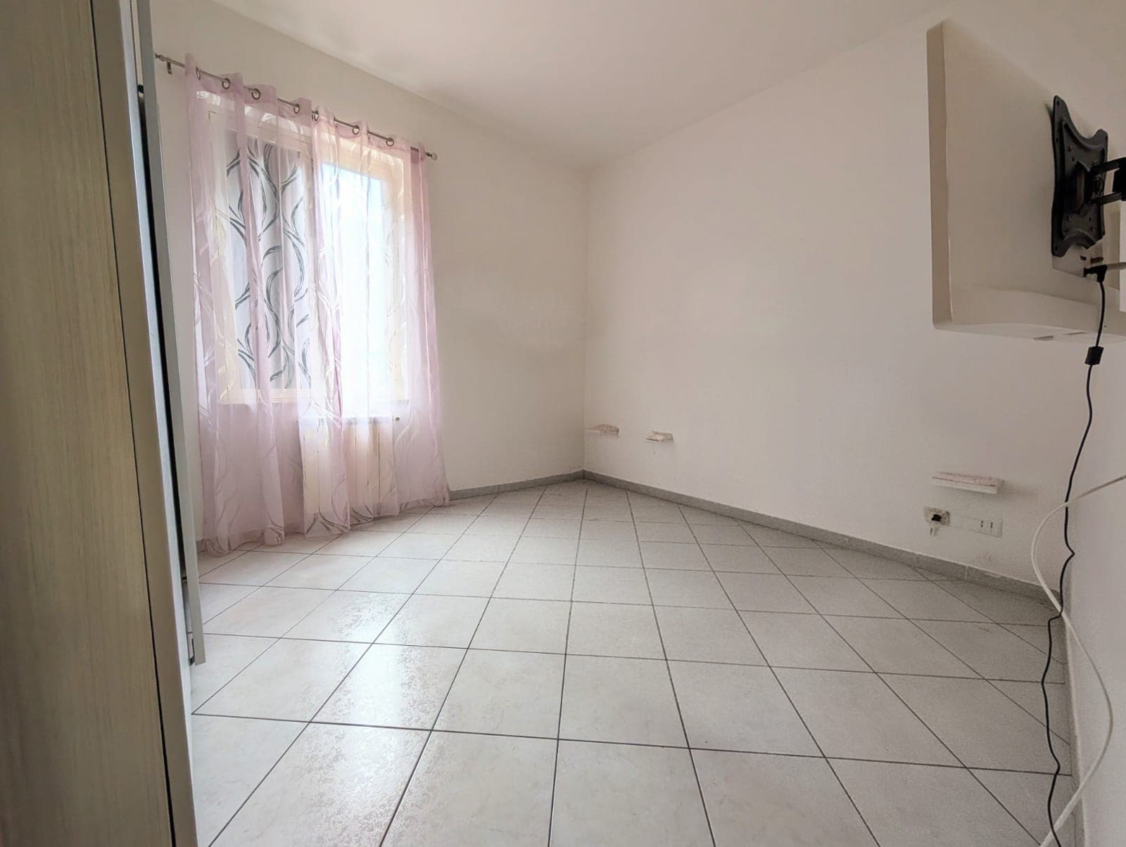 Ampio soggiorno vuoto con pareti bianche, finestra con tende rosa e pavimento in piastrelle chiare, ideale per arredamento moderno e luminoso.