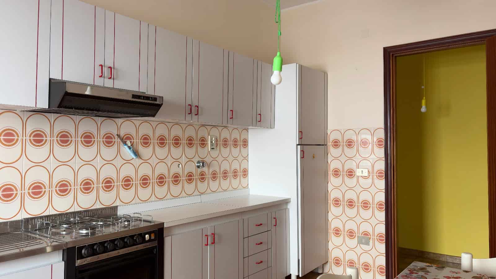 Spazio cucina moderno con mobili bianchi, dettagli rossi e pareti rivestite con piastrelle decorative a motivi geometrici in toni caldi, ideale per ambienti abitativi funzionali e di design.