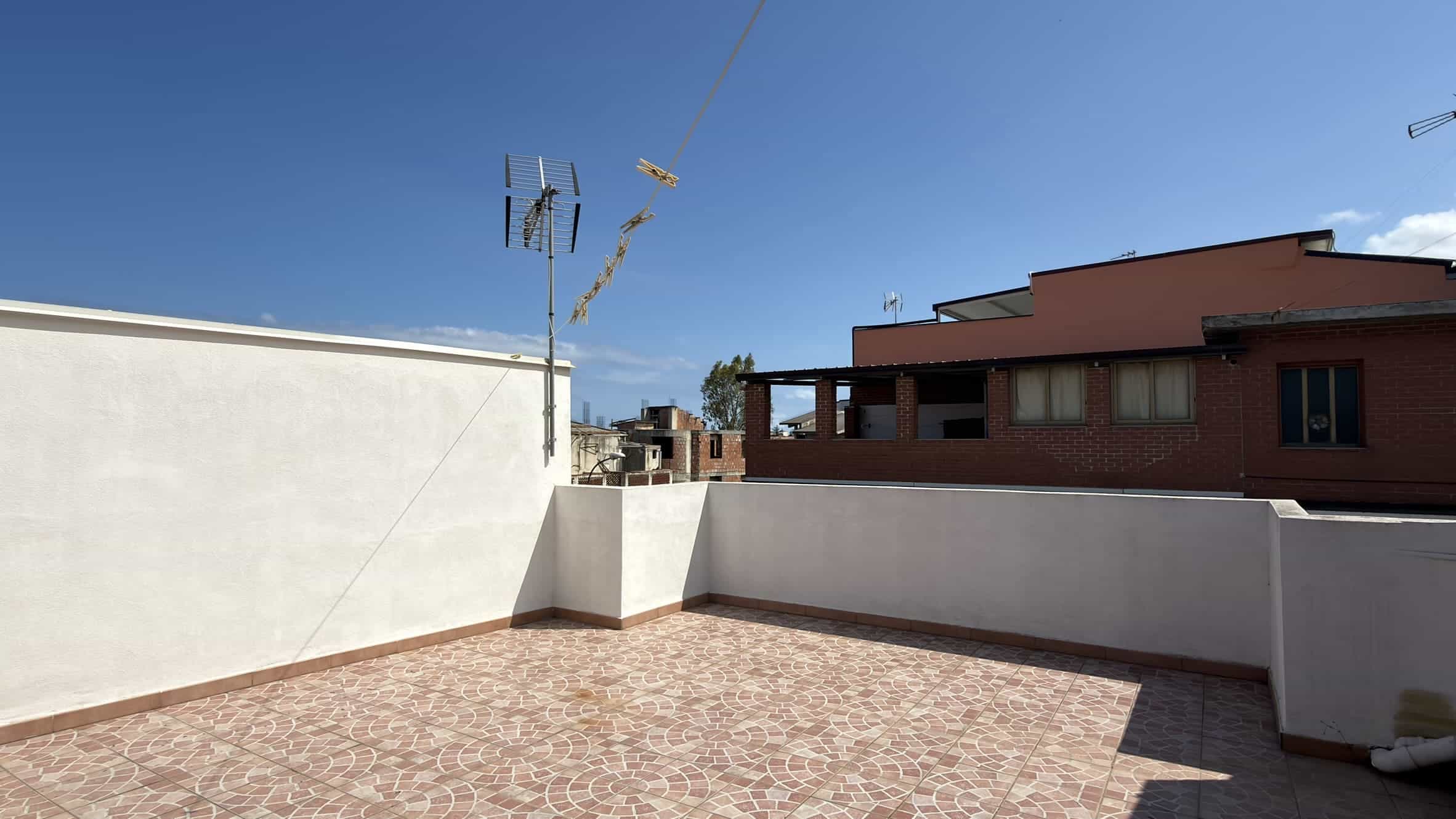 Ampio terrazzo all'aperto con pavimentazione in piastrelle, vista sul cielo azzurro e su edifici circostanti, ideale per relax e socializzazione.
