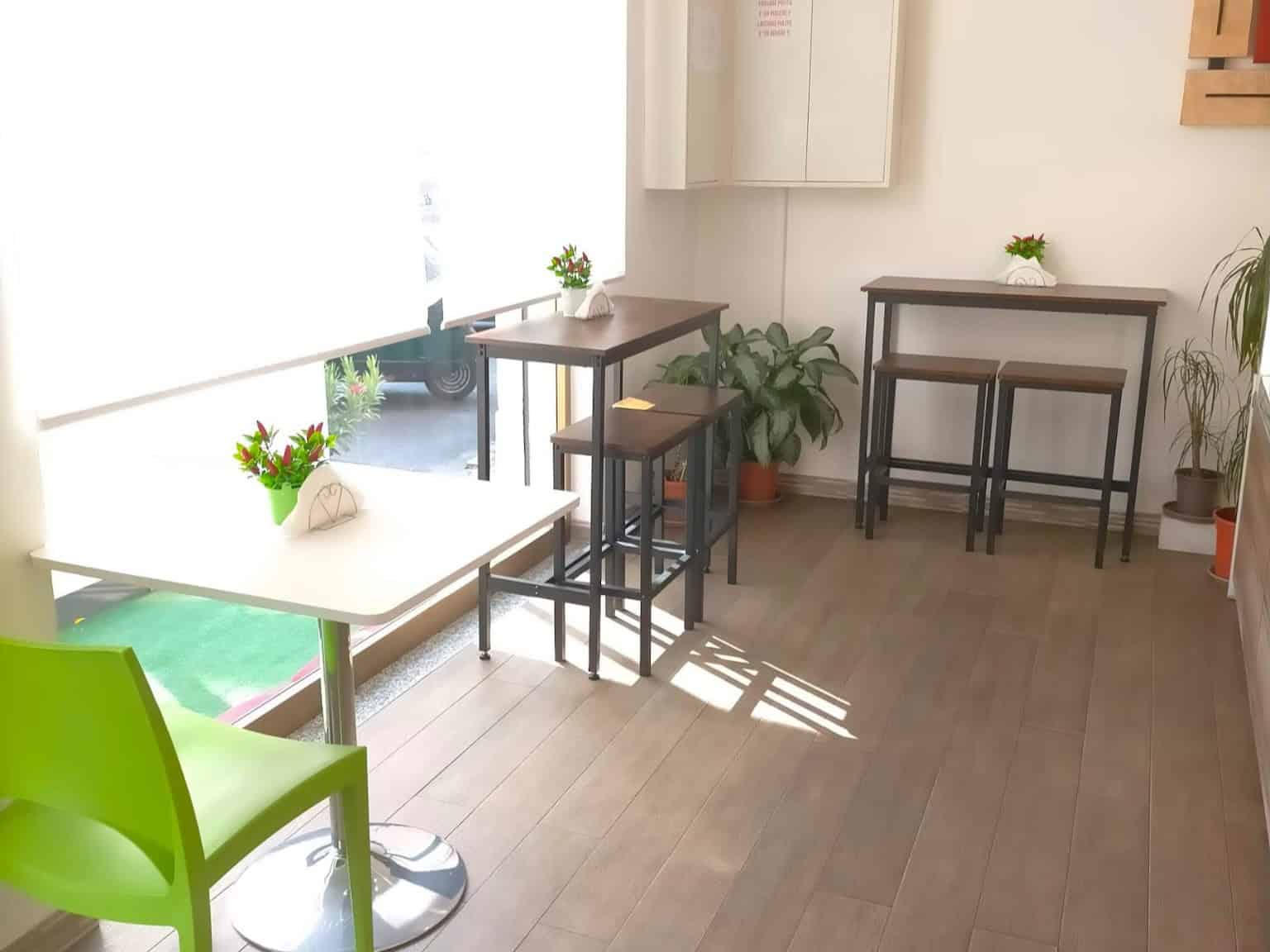 Panca alta in legno con sgabelli in un ambiente luminoso e accogliente, decorato con piante verdi e fiori, ideale per caffè o spazi di lavoro informali, in stile moderno e minimalista.