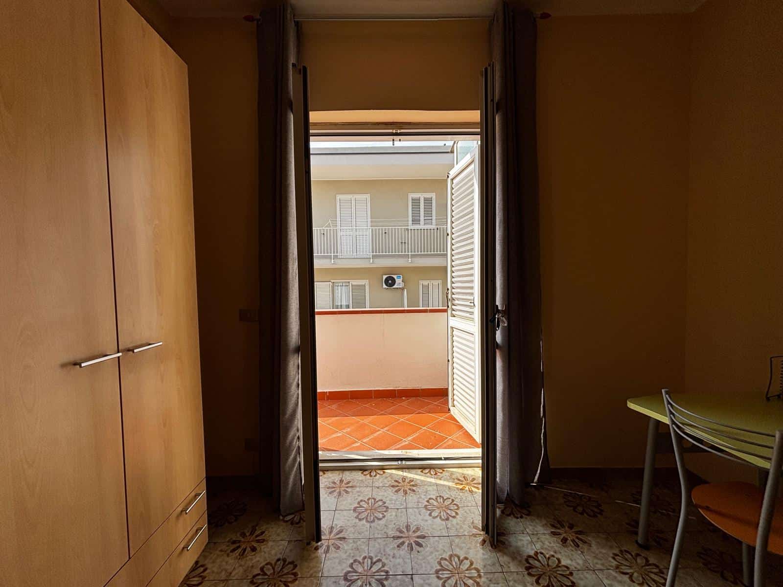 Porta finestra che si apre su un balcone con pavimentazione in piastrelle arancioni, all’interno di una stanza con armadio in legno e pareti beige.