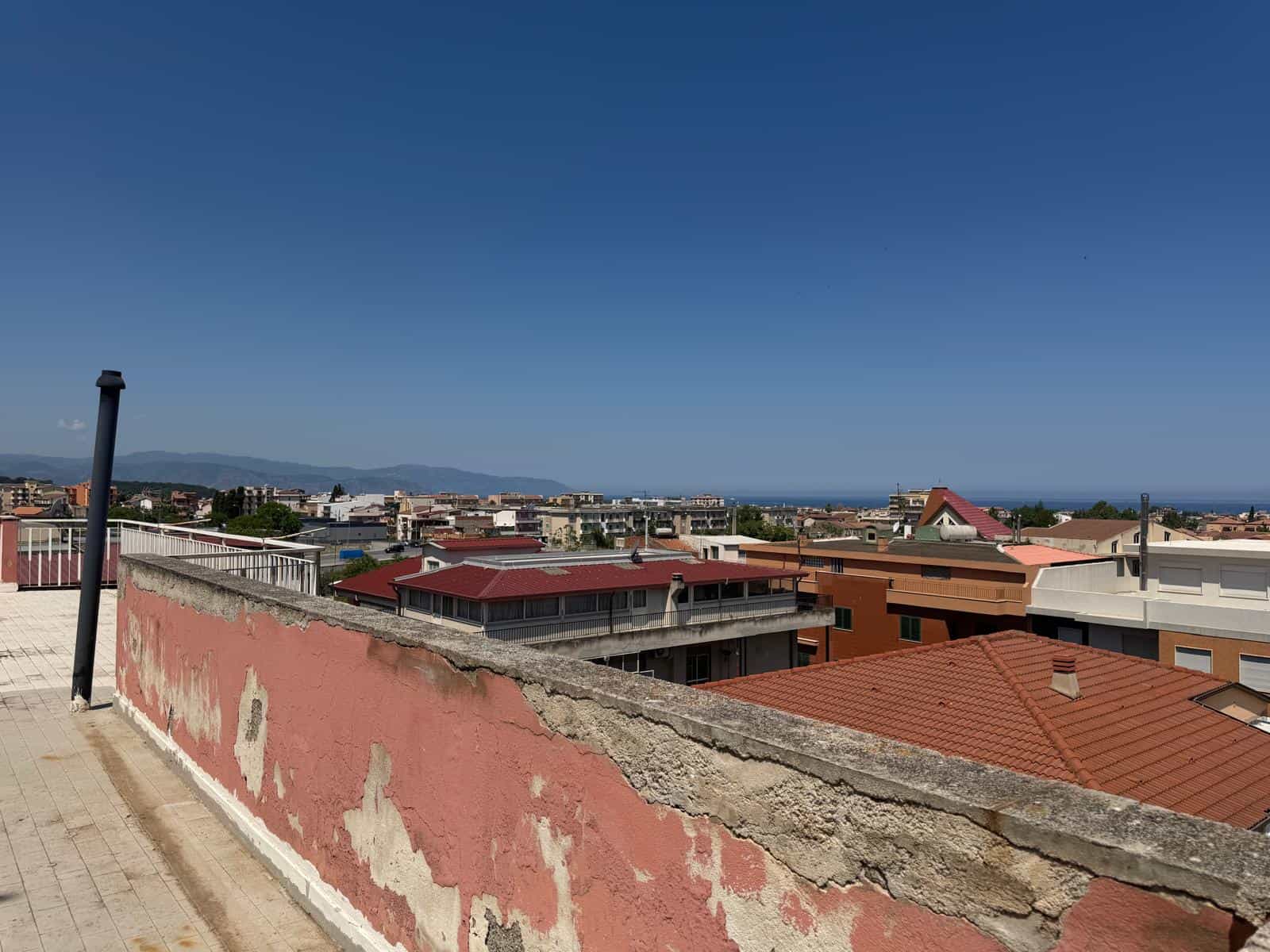 Balcone con vista panoramica sulla città e sul mare, in un contesto urbano residenziale, con tetti rossi e edifici moderni, sotto un cielo sereno, ideale per immobili e proprietà in vendita o affitto.