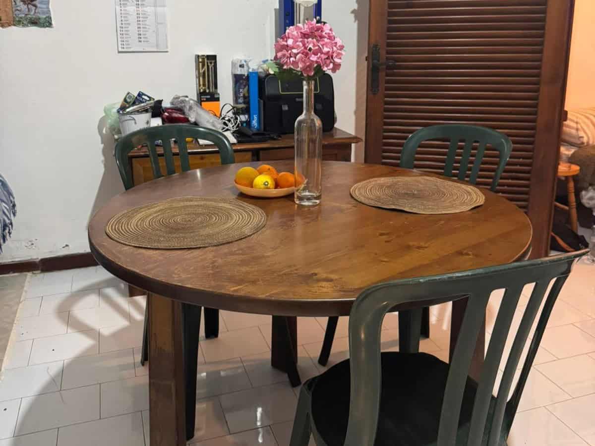 Tavolo da pranzo rotondo in legno con fiori rosa in vaso e frutta fresca, ambiente domestico accogliente e ben arredato.